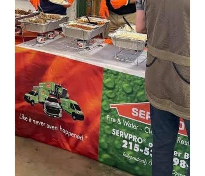 Three people at a serving table with trays of meat. The table cloth is orange and green with SERVPRO's logo on it.